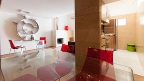 Un salon moderne avec une table à manger en verre, des chaises rouges, une grande horloge murale et un coin cuisine équipé d’appareils électroménagers en acier inoxydable.