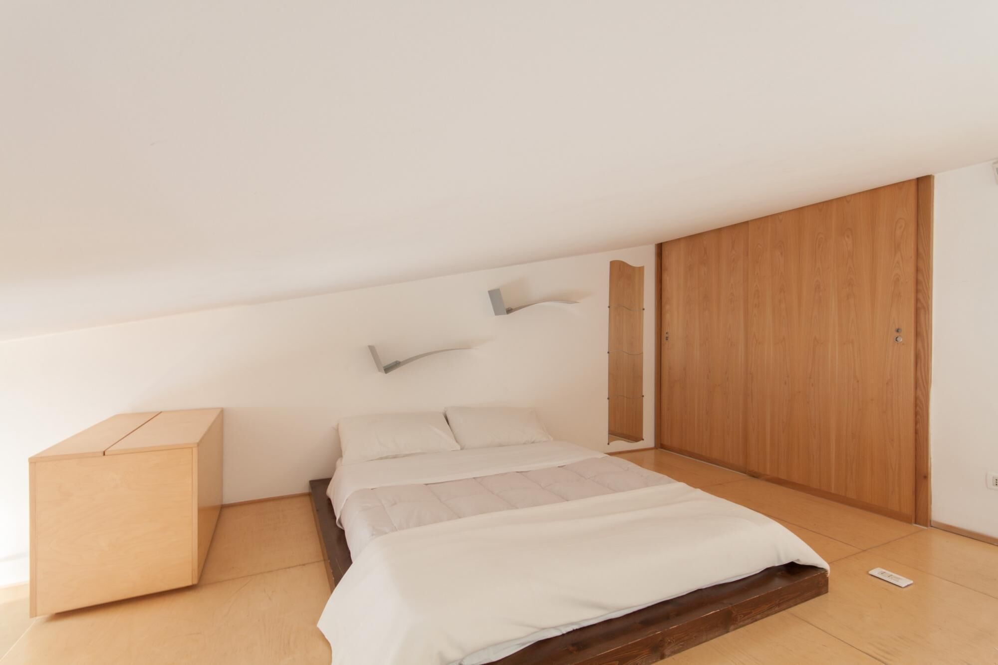 Quarto minimalista com cama, guarda-roupa de madeira e um pequeno armário.