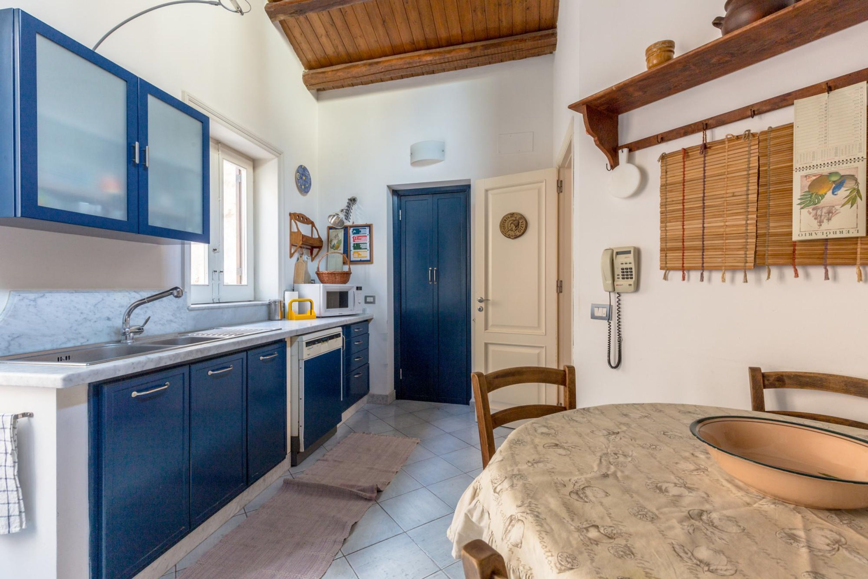 A kitchen with blue cabinets, a white countertop, a wooden ceiling, and a dining table with a glass bowl.
