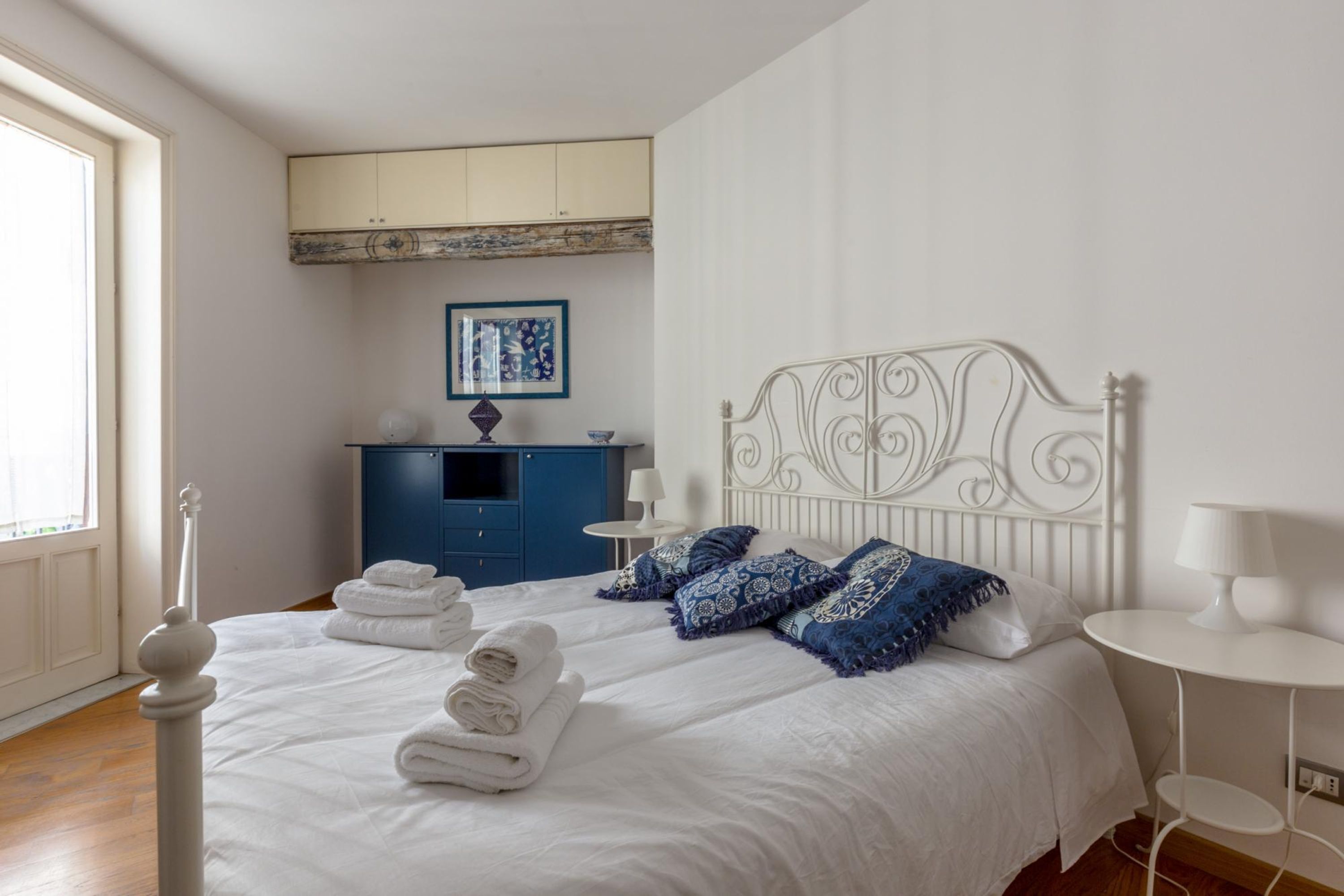 Un llit ben fet amb llençols blancs i coixins blaus estampats, una petita taula de nit amb una làmpada, i un prestatge blau amb un quadre emmarcat a la part superior.