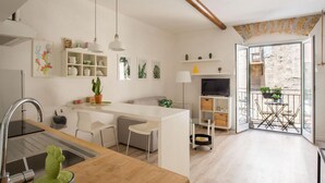 Una cocina moderna con armarios blancos, encimera de madera y una isla integrada. Hay un comedor con mesa y sillas, un televisor y un balcón con plantas en macetas.