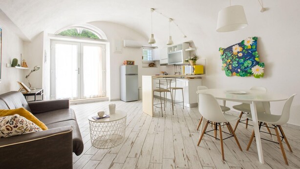 Un vaste espace de vie comprenant un canapé, une table basse ronde et une table à manger avec des chaises. On aperçoit au fond une cuisine avec des meubles blancs et un réfrigérateur.