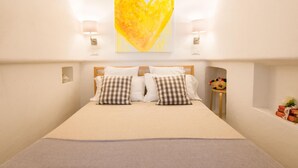 Une chambre avec un grand lit, deux lampes de chevet, une œuvre d’art en forme de cœur accrochée au mur, et une petite table sur laquelle se trouvent un panier et des livres.