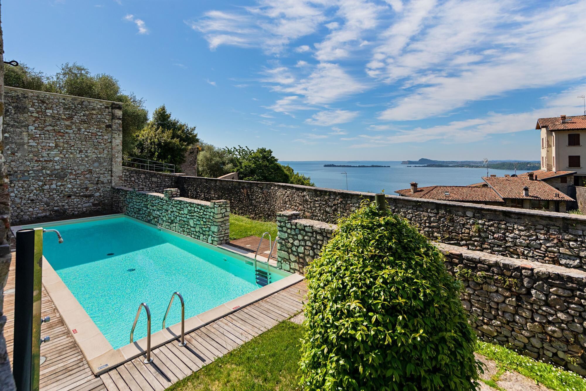 A stone-walled pool with a wooden deck, surrounded by greenery and overlooking a body of water with distant landforms.
