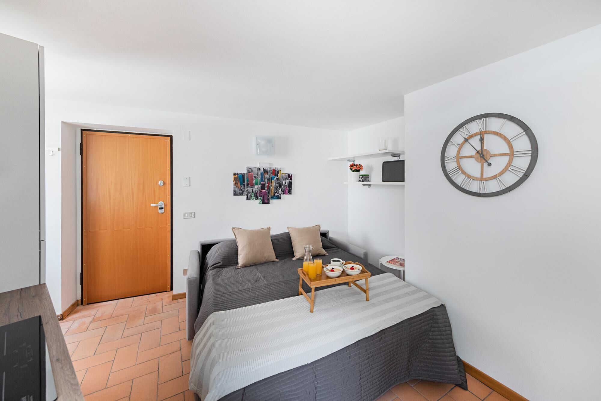 A bedroom with a bed, a wooden door, a wall clock, and a small table with breakfast items.