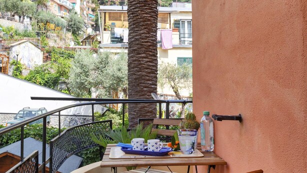 A balcony with a table set for two, a palm tree, and a view of a hillside with buildings.