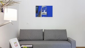 A grey sofa with two cushions, a white lamp, a framed picture, a small table with a green apple and a brown mug, and a blue wall art piece.