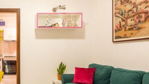 Ett vardagsrum med en grön soffa, ett litet vitt sidobord med en växt och en rosa hylla med dekorativa föremål.