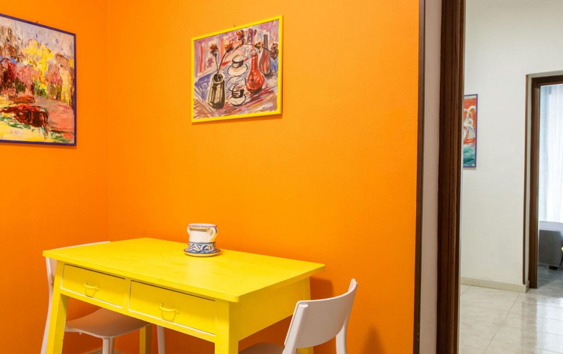 Een kamer met een gele tafel en stoelen, een schilderij aan de muur, en een deur naar een andere kamer.