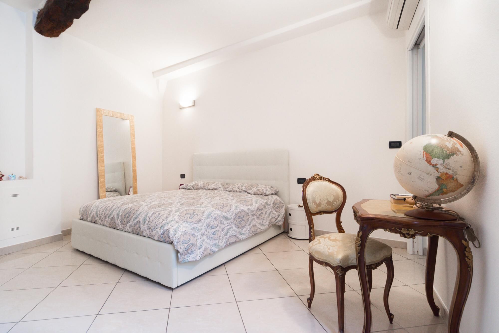 A bedroom with a large bed, a wooden chair, a small table with a globe, and a mirror on the wall.