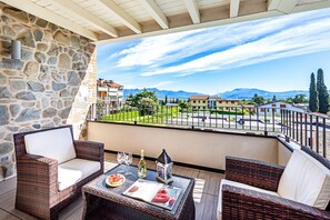 A balcony with wicker furniture, a table with a bottle and glasses, and a view of a town and mountains.