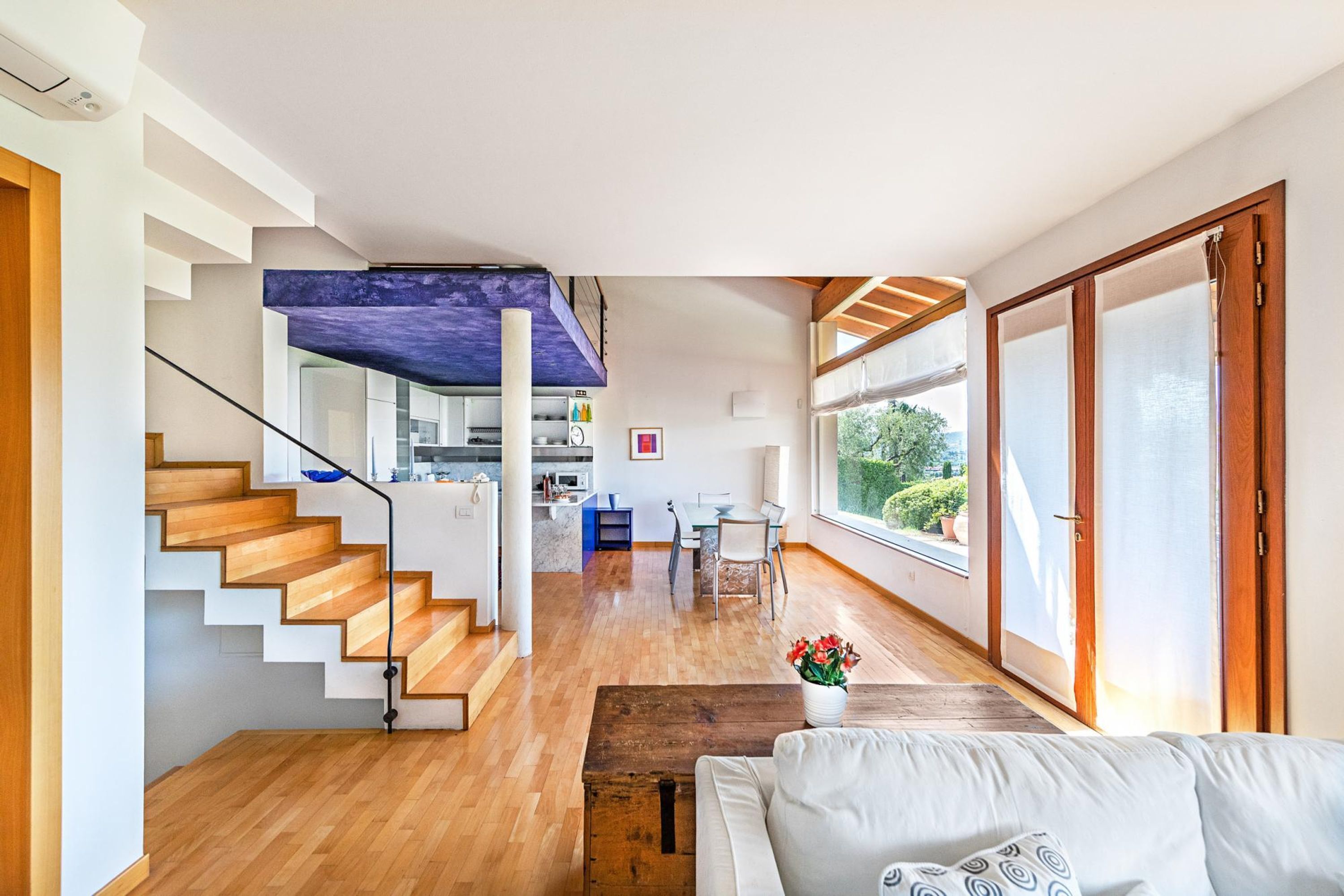 A modern interior with a staircase leading to a loft area, a dining area, and large windows offering a view of the outdoors.
