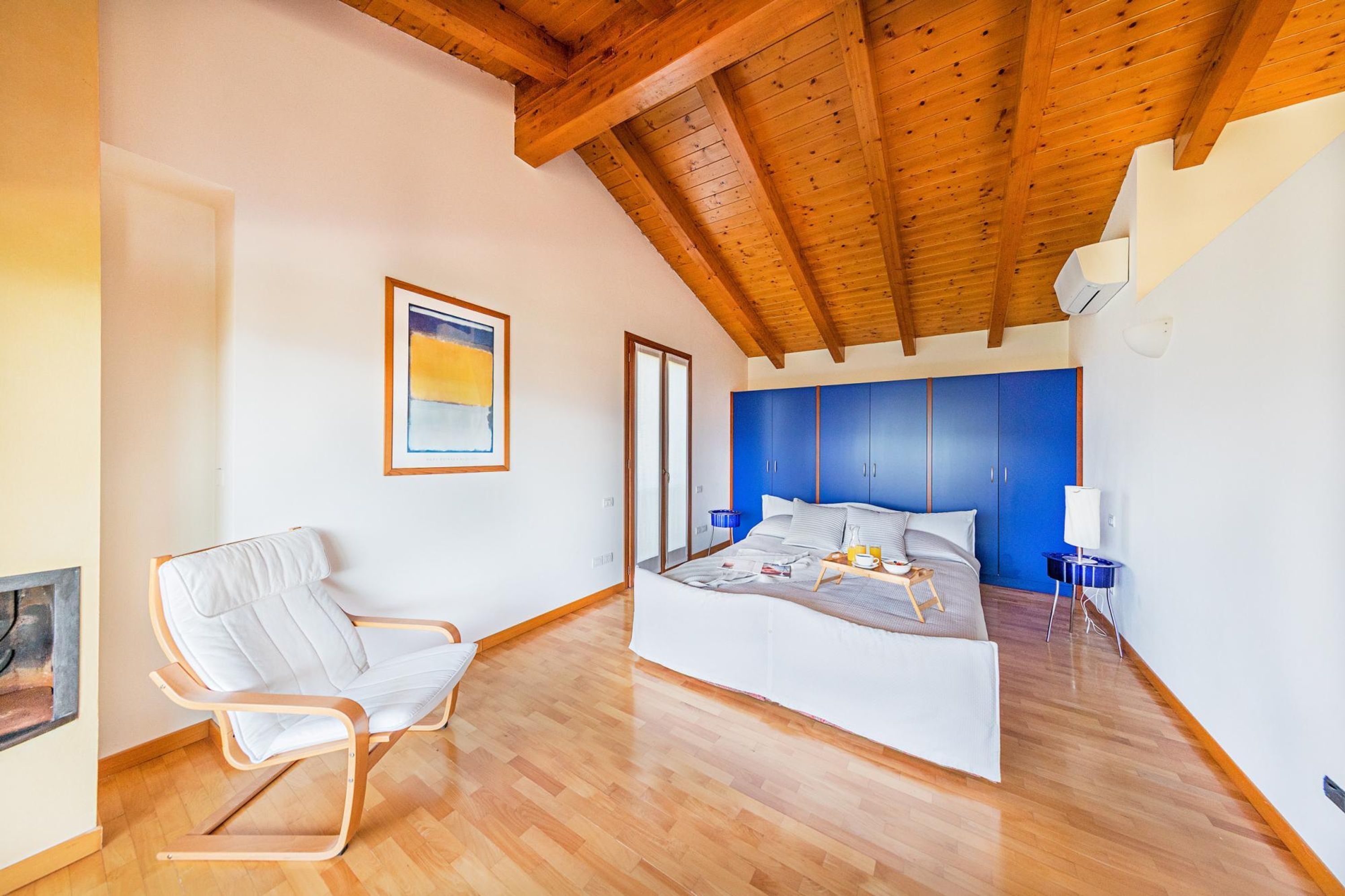 A modern bedroom with a wooden ceiling, a large bed with a blue headboard, a white chair, and a painting on the wall.