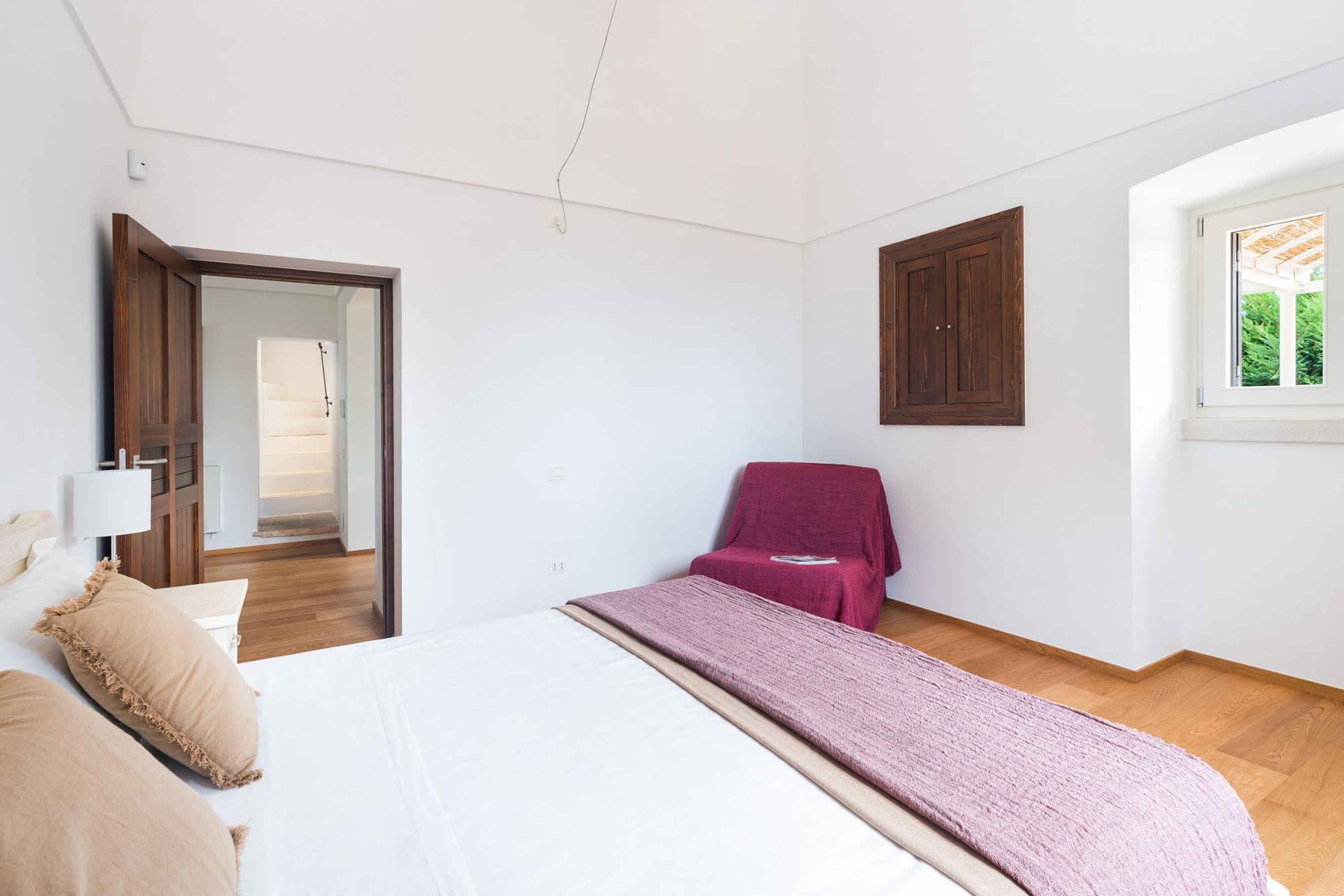 A bedroom with a bed, a chair, a wooden wardrobe, and a window.