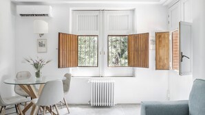 A modern living room with a glass dining table, white chairs, a blue sofa, and a large window with wooden shutters. - Euphorbia Apartment by Wonderful Italy (Catania)