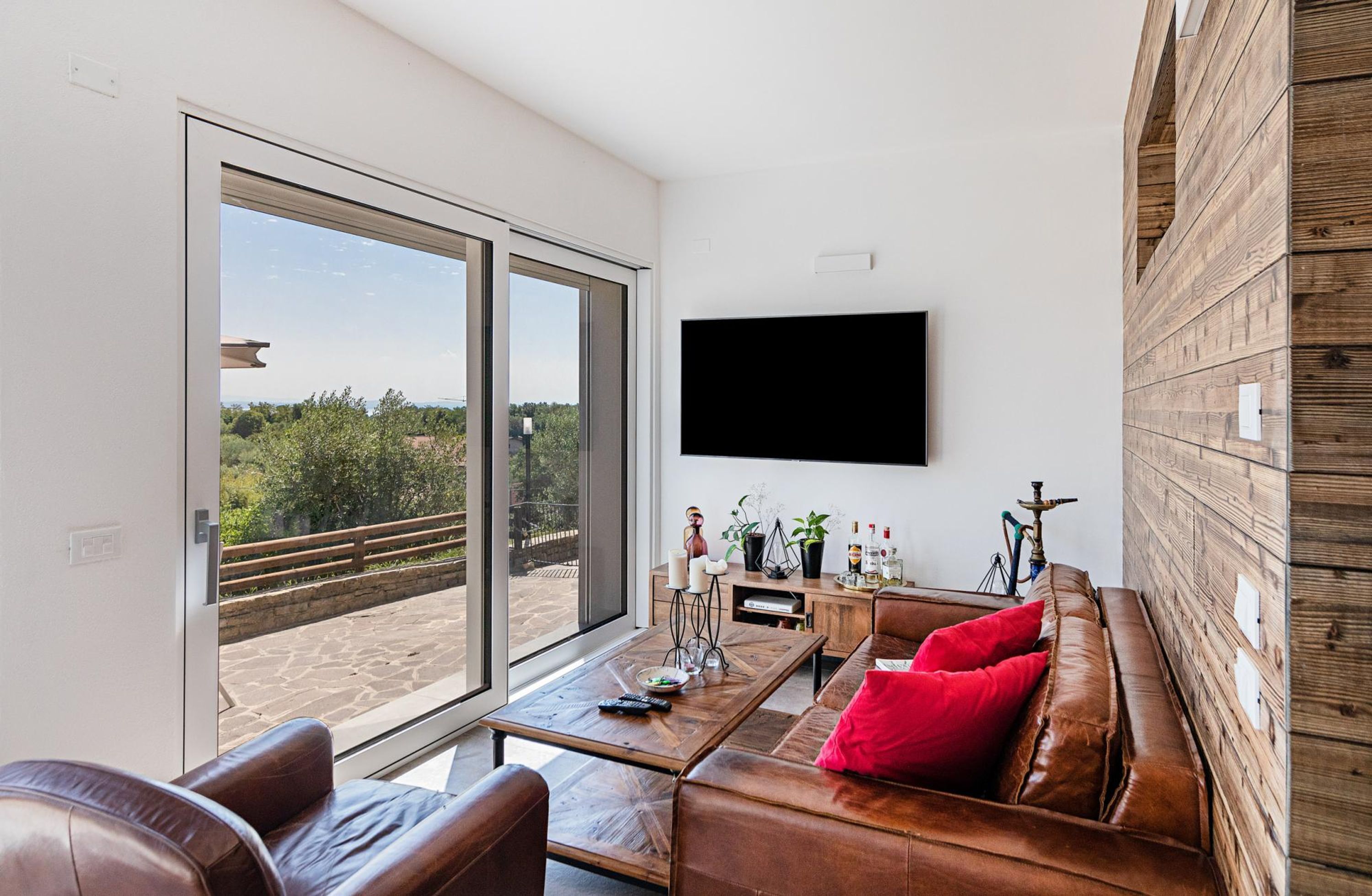A modern living room with a large sliding glass door leading to a balcony, a flat-screen TV mounted on the wall, a wooden coffee table, leather sofas, and a potted plant.