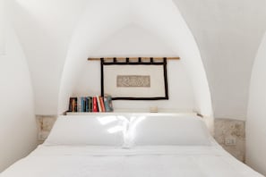 A bedroom with a white bed, a wooden shelf with books, and a decorative wall hanging. - Casa Graziella con Terrazza by Wonderful Italy (Ostuni)