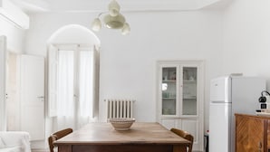 Una habitación luminosa y blanca con una mesa de comedor de madera, un radiador, un armario con puertas de cristal y un refrigerador blanco.