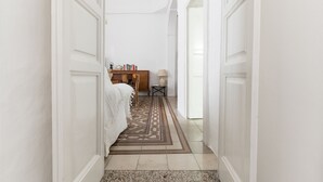 Un pasillo con un tapete estampado, un escritorio de madera con libros y una lámpara, y una cama con ropa de cama blanca.
