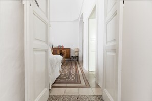 Un corridoio con un tappeto decorato, una scrivania in legno con libri e una lampada, e un letto con lenzuola bianche.