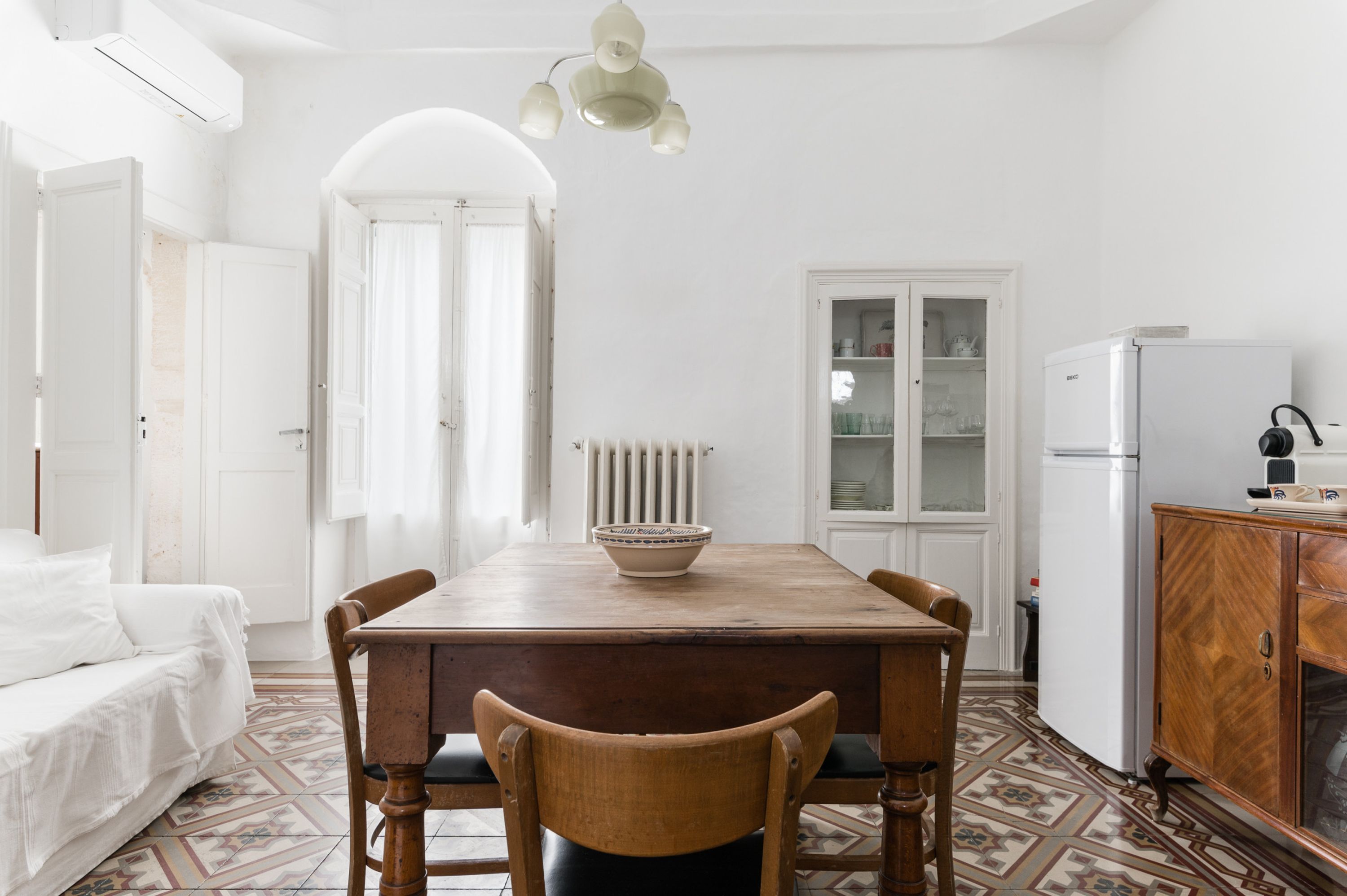 A white room with a wooden dining table, chairs, a white sofa, a radiator, a cabinet, and a window with curtains.