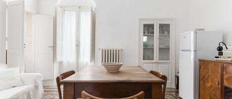 A white room with a wooden dining table, chairs, a white sofa, a radiator, a cabinet, and a window with curtains.