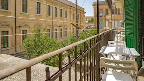 A balcony with a metal railing, white plastic chairs, and a view of yellow buildings with green shutters. - Cavour Apartment Near the Cathedral by Wonderful Italy (Noto)
