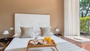 A hotel room with a large bed, two pillows, a bedside table with a lamp, a tray with breakfast, and a window with a view of greenery.