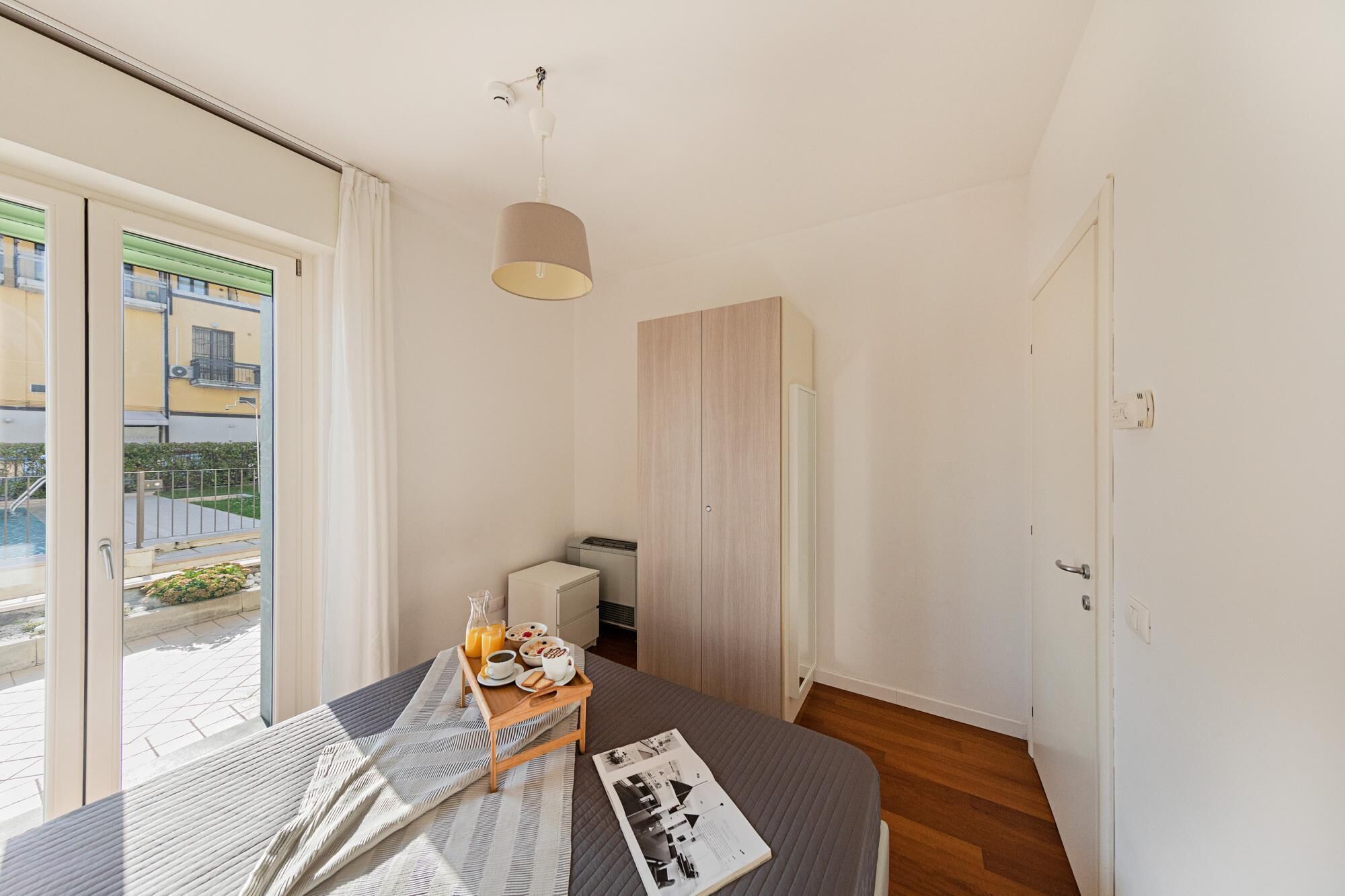 Une chambre à coucher avec un lit, une table de chevet sur laquelle se trouvent des éléments pour le petit-déjeuner, une armoire et une vue sur un bâtiment à l’extérieur.