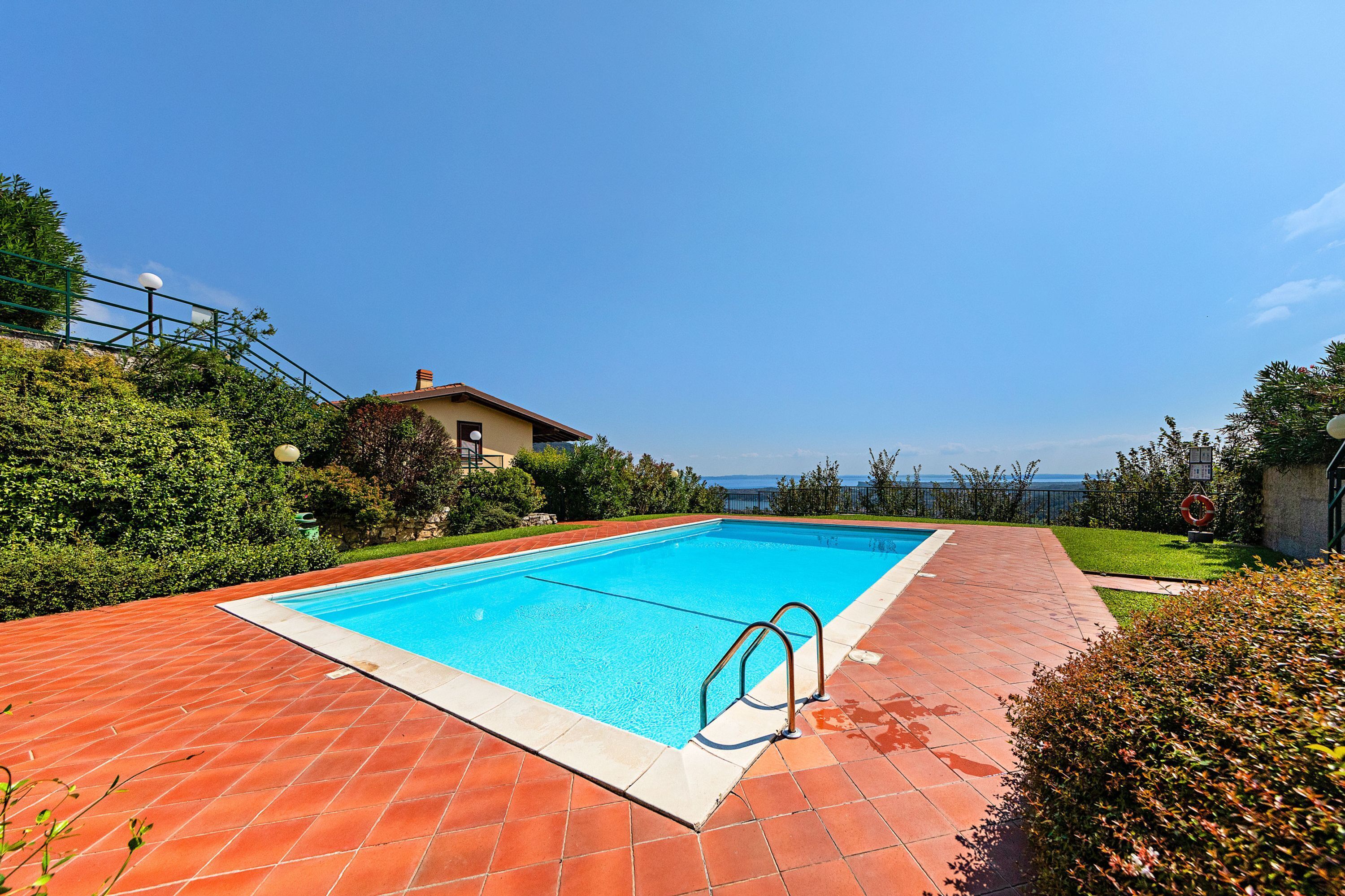 A rectangular swimming pool with a tiled deck, surrounded by greenery and a clear view of the sea.
