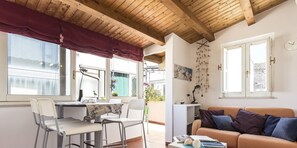 Une pièce chaleureuse avec un plafond en bois, un coin repas avec des chaises et une table, un canapé avec des coussins et une fenêtre donnant sur un paysage verdoyant.