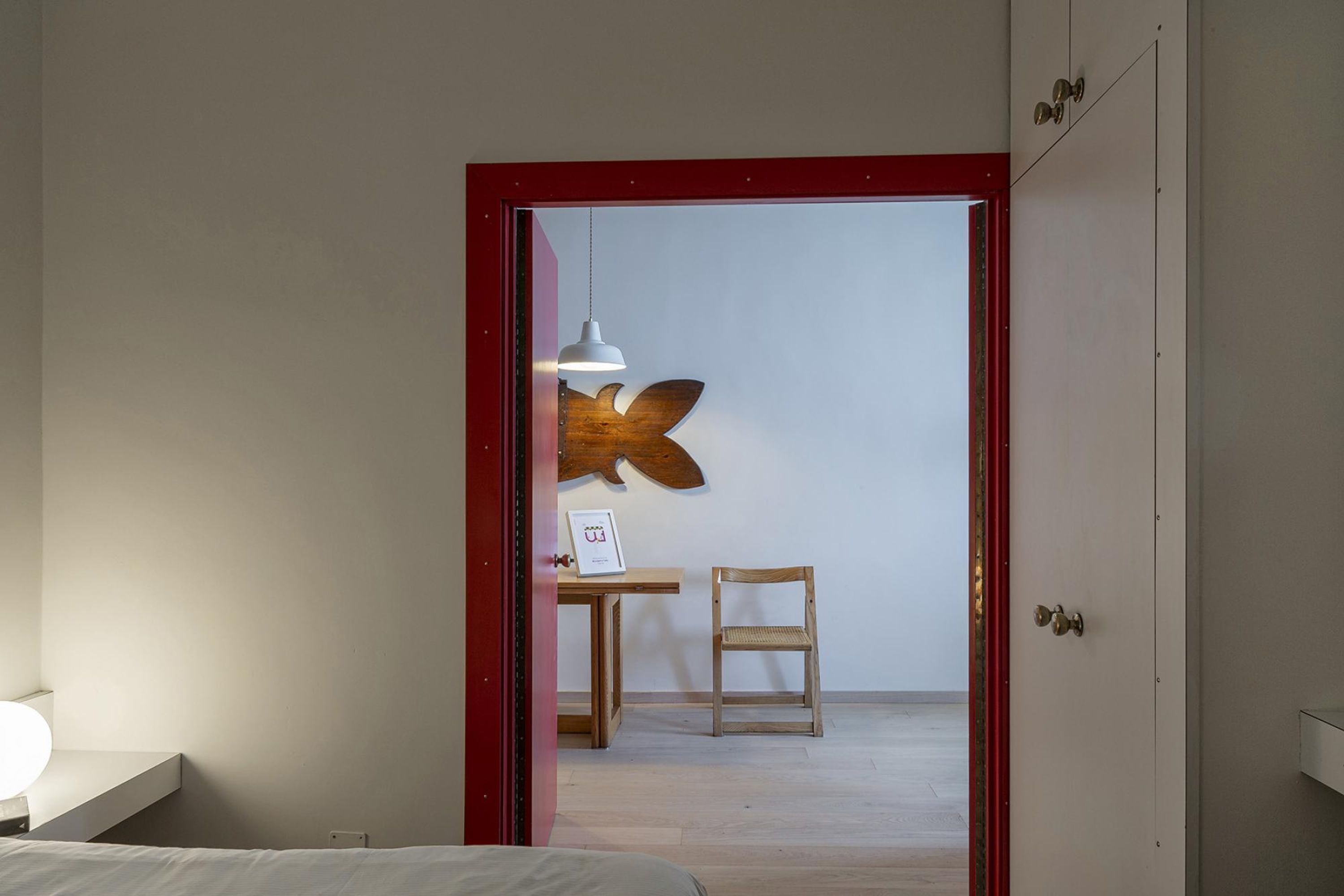 Een hotelkamer met een bed, een bureau met stoel, een wandlamp en een deur met een rode omlijsting.