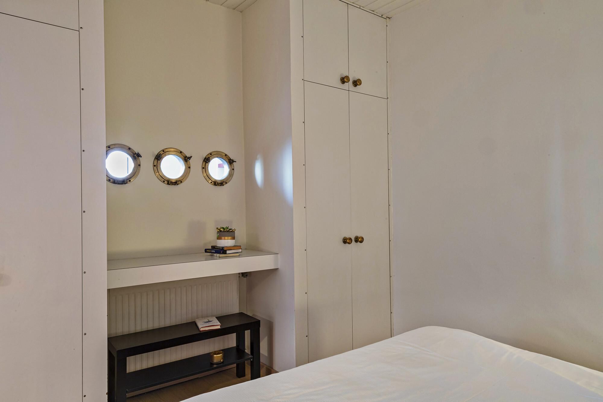 Um quarto com cama, mesa de cabeceira, estante com livros e planta, e guarda-roupa com espelho e porta.