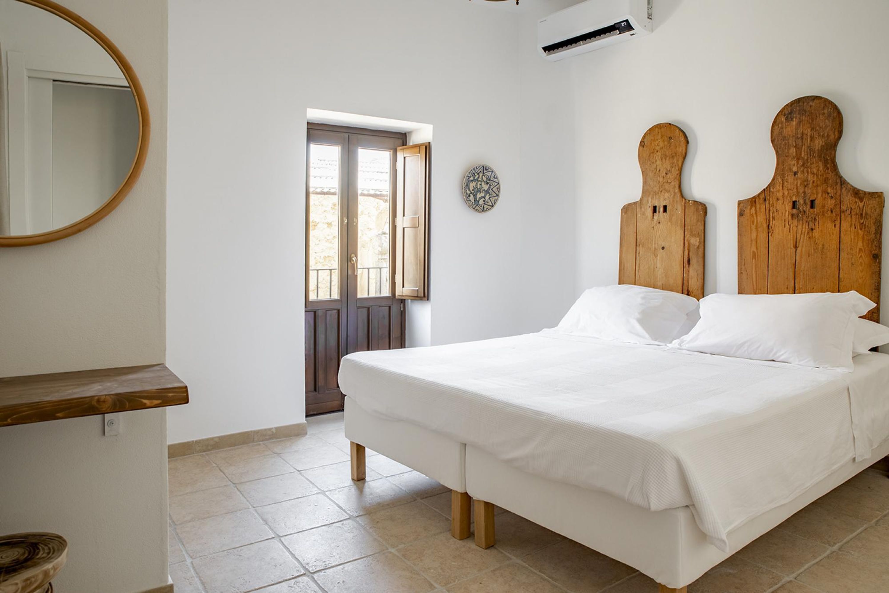 A bedroom with a wooden bed, white bedding, a round mirror, and a door leading outside.
