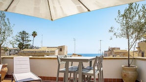 Eine Terrasse mit Tisch, Stühlen, einer Pflanze im Topf und Blick aufs Meer.