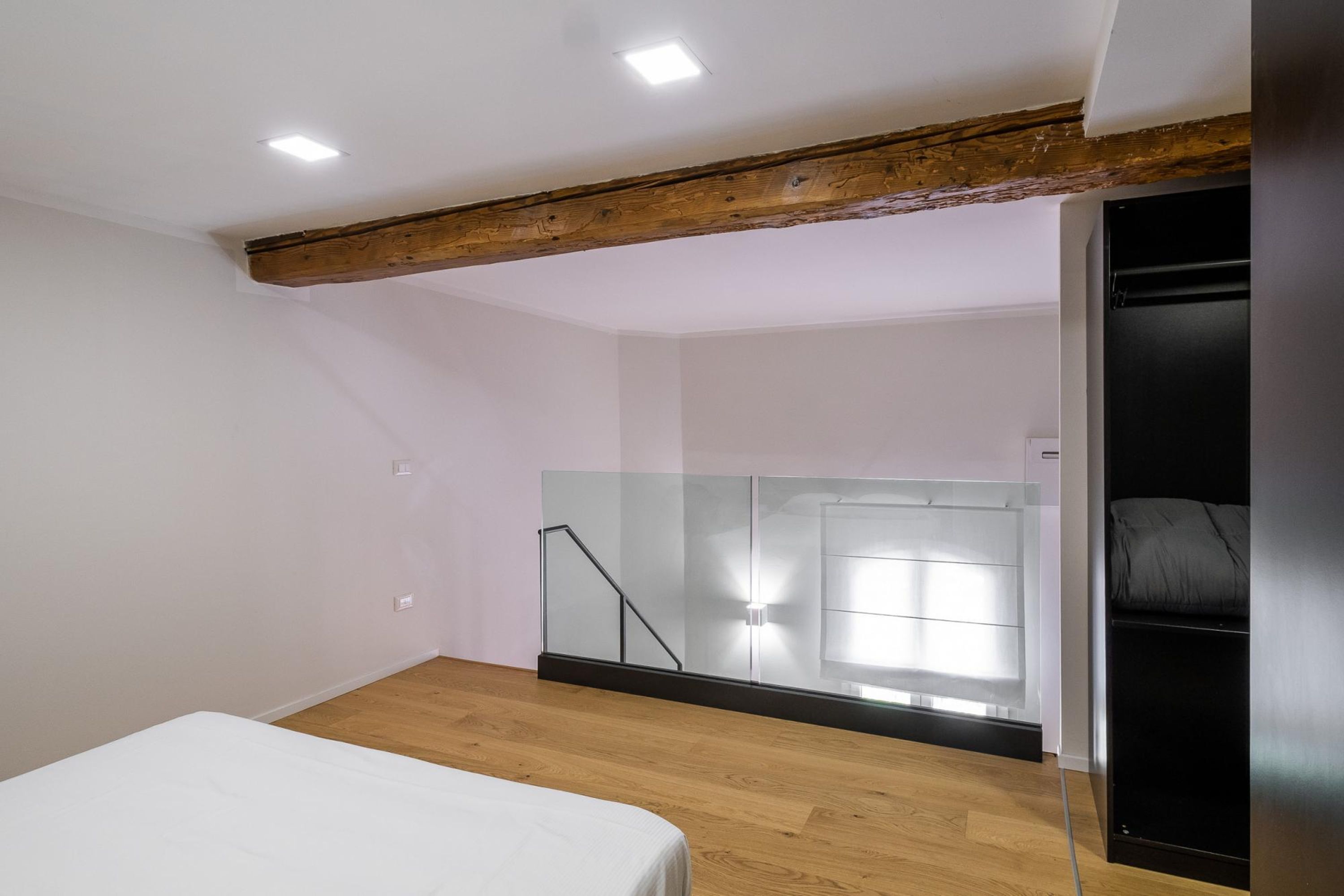 A bedroom with a wooden ceiling beam, a bed, a black wardrobe, and a glass partition with a staircase.