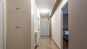 Un pasillo estrecho con un radiador, una puerta al final y una cama visible a través de una puerta abierta.