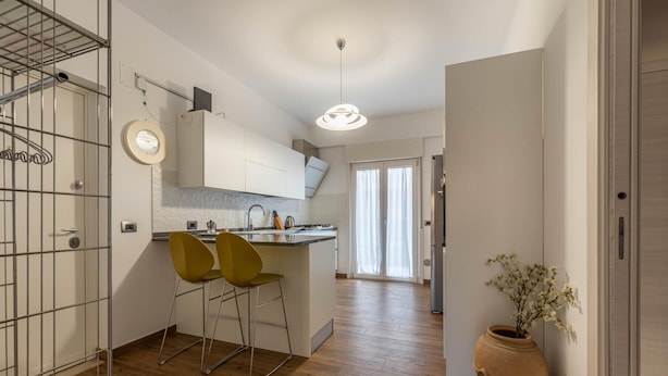 Una cocina moderna con una isla central, dos sillas amarillas, un ventanal grande y una planta en maceta.