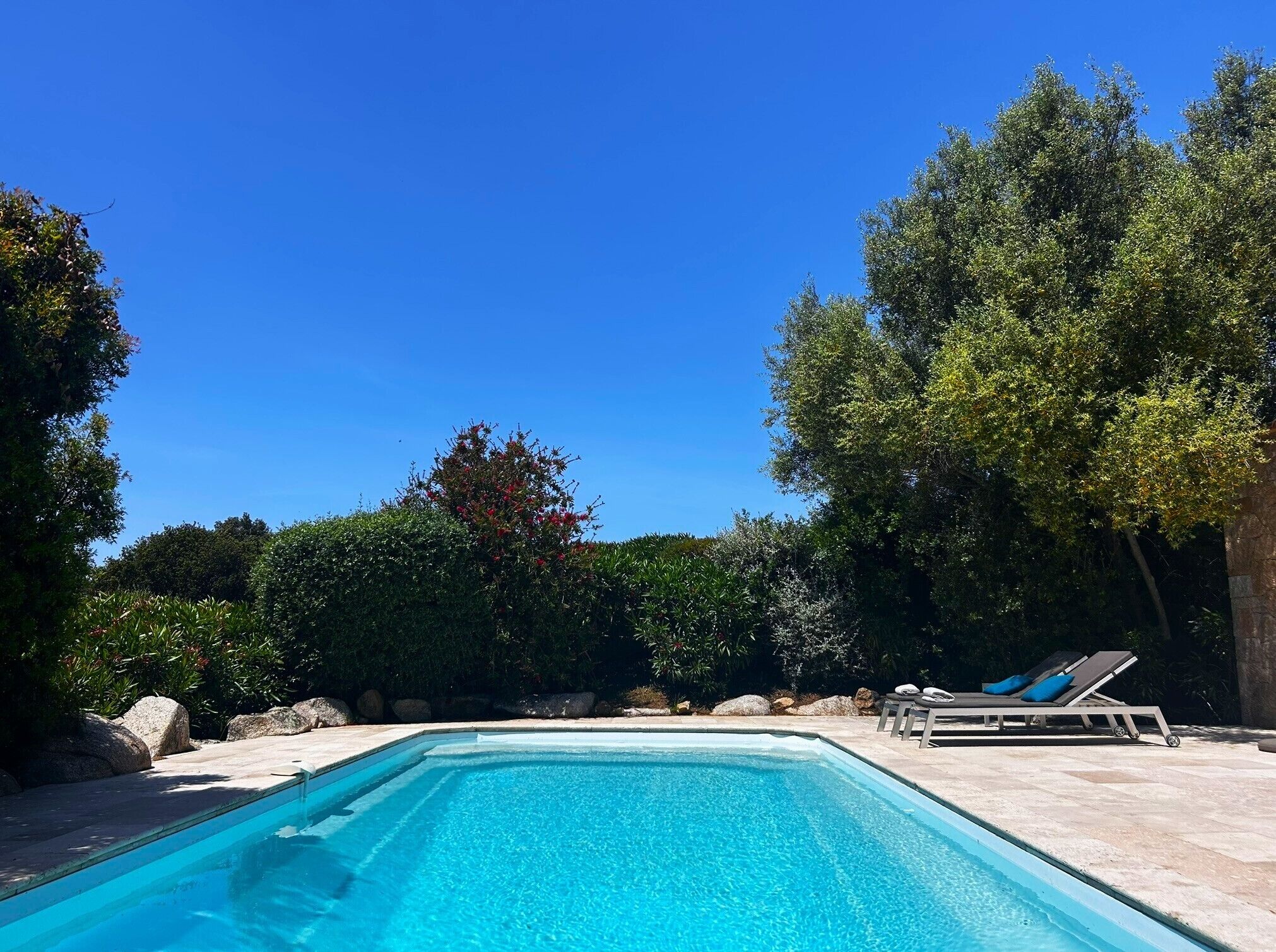 Una piscina rectangular con agua azul cristalina, rodeada por una terraza de piedra. Se observan sillones de descanso con cojines a la derecha.