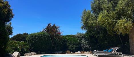 Una piscina rectangular con agua azul cristalina, rodeada por una terraza de piedra. Se observan sillones de descanso con cojines a la derecha.