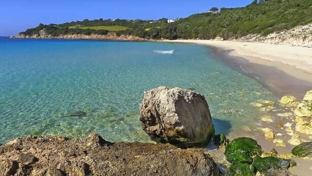 Une plage côtière aux eaux cristallines, avec un grand rocher au premier plan et une rive sablonneuse.