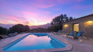 Une piscine avec des chaises longues et un bâtiment en pierre illuminé, entouré de rochers et d’arbres, au coucher du soleil.
