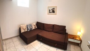 Ein Eckwohnzimmer mit einem braunen Modulsessel, einem kleinen Tisch mit Lampe, einem gerahmten Bild an der Wand und einem Fenster mit Jalousien.