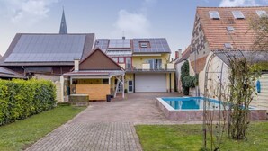 Un quartiere residenziale con un vialetto lastricato che porta a una casa con garage, piscina e il campanile di una chiesa sullo sfondo.