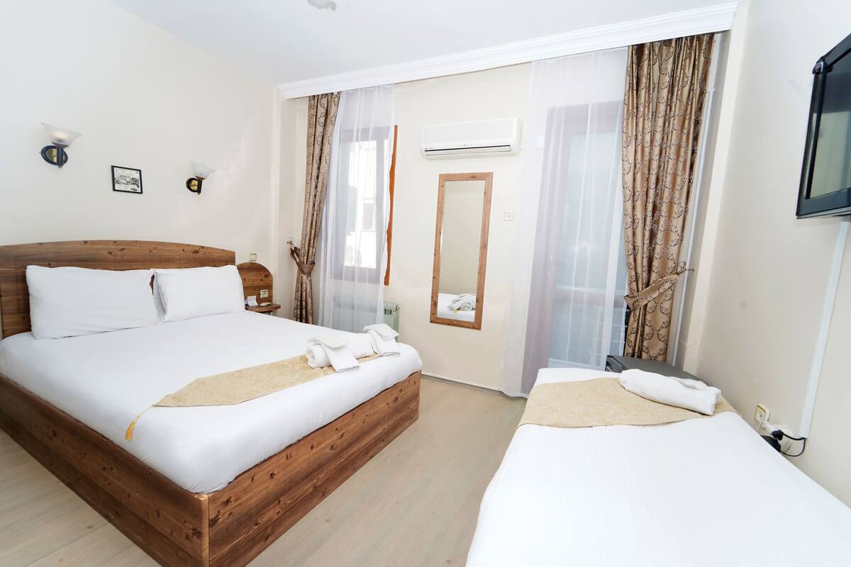 Hotelkamer met een houten bed, een bijbed, een spiegel, een televisie en een raam met gordijnen.