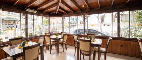 Un restaurante con mesas y sillas de madera, ventanas amplias y piso de baldosas.