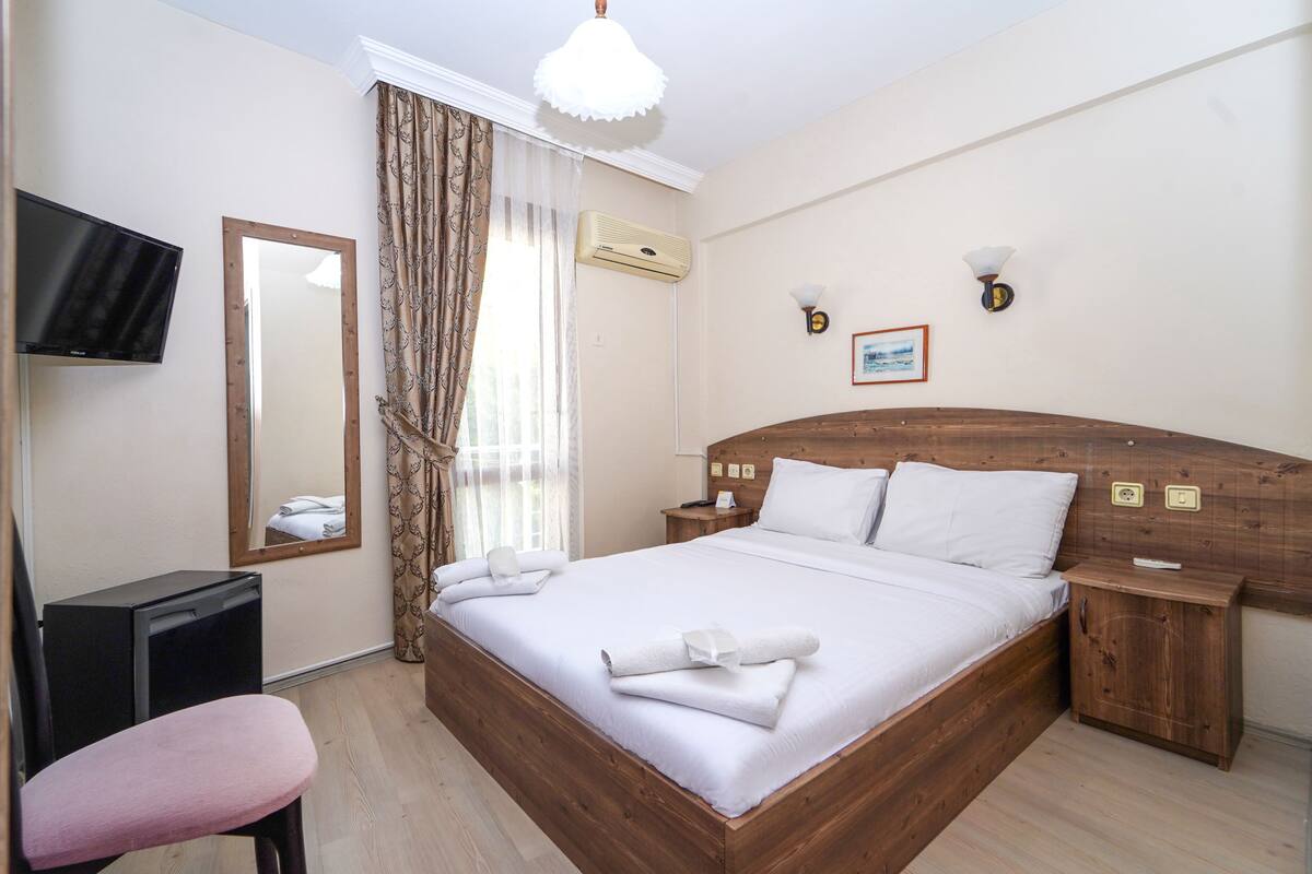 Een keurig opgemaakt bed met witte lakens, een houten hoofdbord, een nachtkastje, een klein houten kastje, een aan de muur bevestigde televisie en een raam met gordijnen.