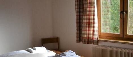 Quarto com duas camas de solteiro, cabeceira de madeira, janela com cortinas xadrez e radiador.
