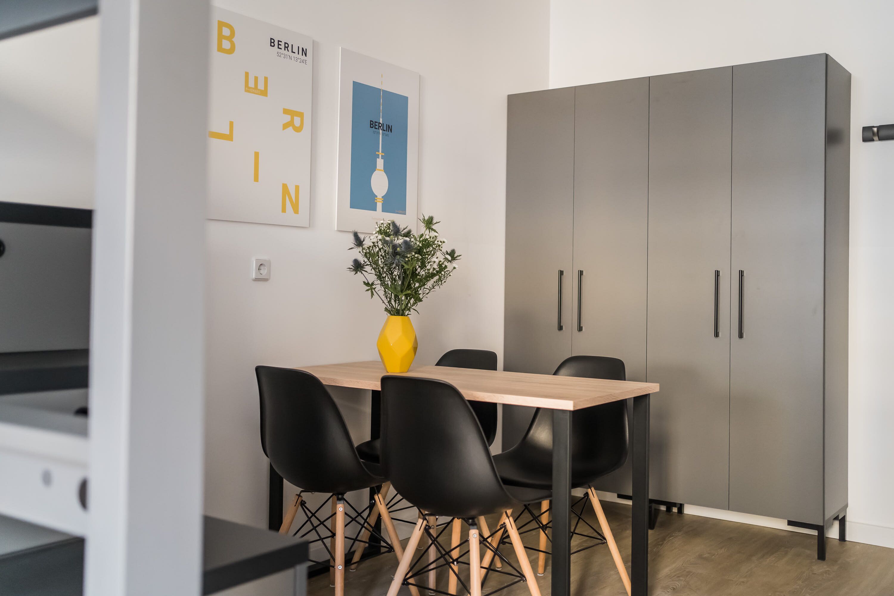 A modern dining area with a wooden table, black chairs, and a yellow vase with green plants.