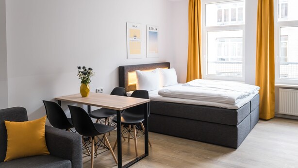 Une chambre d’hôtel moderne, équipée d’un lit, d’un bureau avec des chaises, d’un vase avec des fleurs et de deux tableaux encadrés accrochés au mur.